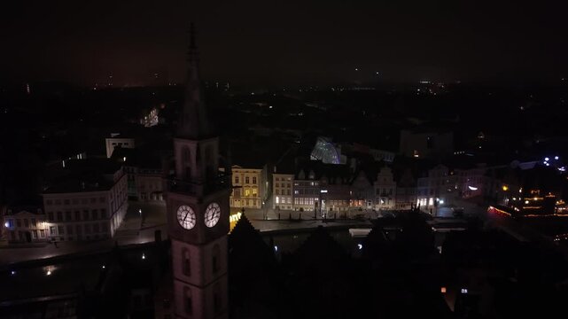 Night aerial orbit of the historic Ghent Belfry clock tower, revealing the glowing medieval cityscape and the scenic Leie river canal in Ghent, Belgium.
