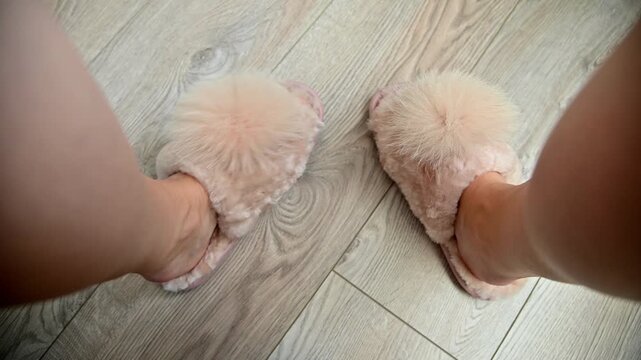 Female feet in fluffy pink slippers with pom-poms positioned on light wooden floor, showcasing a cozy indoor setting with soft textures and warm tones