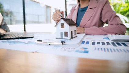 A businesswoman reviews financial charts and documents beside a small house model, analyzing real estate investment and mortgage planning at her desk. © Natthawadee