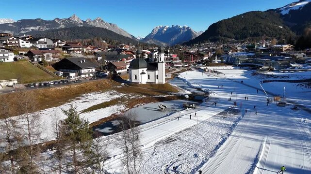 perspective looks upon iconic white walled seekirchl church cross country skiers glide across meticulously groomed snow tracks wind through sunlit seefeld valley down drone tirol austria 