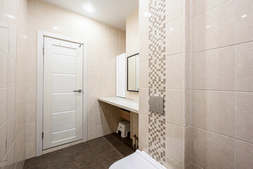 Neutrally-toned bathroom with beige tiles, wall-mounted toilet, built-in sink with mirror, recessed ceiling lights, white door, mosaic tile accent strip, small step stool