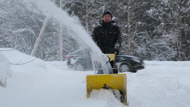 Slow motion 4K footage of a man clearing the driveway to the road with the snow blower during heavy snowfall, cars passing by. Cold weather, winter maintenance and daily life in rural area