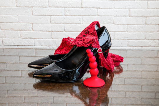 Black high heel shoes with red lace panties and red anal plug on glass table against white brick wall