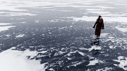 Fototapeta premium Woman in long fur coat walking on dark ice of vast frozen lake winter landscape