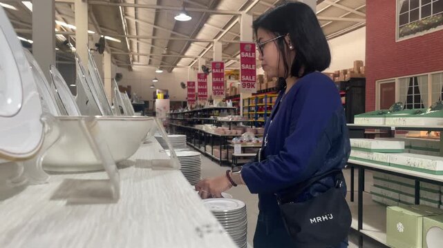 Young Asian woman shopping for ceramic plates in home goods warehouse outlet