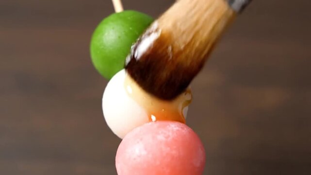 Close-up of a brush applying sauce to colorful dango skewers, a traditional Japanese sweet.