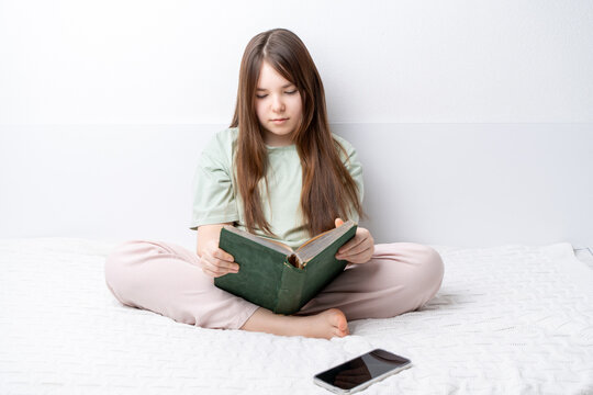 A teenage girl reads a book sitting at home on a bed in a cozy environment, with a phone next to her. The concept of digital detox, analog childhood.