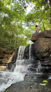 Man sitting on a rock near a waterfall, enjoying a peaceful natural landscape. Concept of slow living, mental wellness, stress relief, digital detox, travel escape and connection with nature.