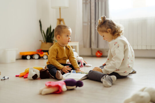 Two kids sit on the floor with toys around them as they read a book together in a well-lit room during daytime playtime
