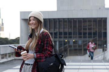 Smiling adult female wearing beanie standing on tiled plaza holding phone and duffel, copy space © wavebreak3