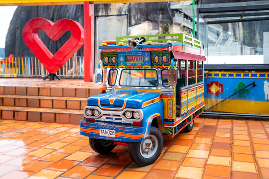 Guatape, Colombia - November 20, 2025: Brightly painted miniature chiva sits on orange tiles near heart sculpture in a tourist zone of Guatape near Medellin, Colombia, during a cloudy day