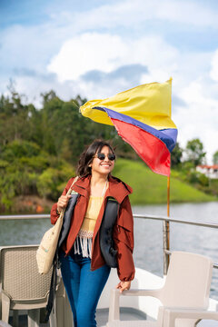 Young happy smiling Latina woman enjoys a peaceful boat tour on the lake once home to Pablo Escobar&rsquo;s house, waving Colombian flag in Guatape near Medellin, Colombia