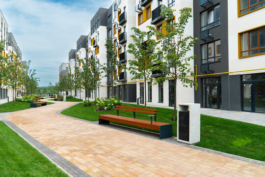 Paved pedestrian path with wooden benches in a modern residential complex courtyard with stylish apartment buildings and green lawn in summer