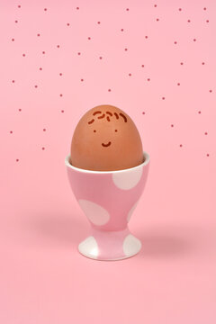 A brown chicken egg with a simple hand drawn smiling face sits in a pink polka dot egg cup against a pink background with dots