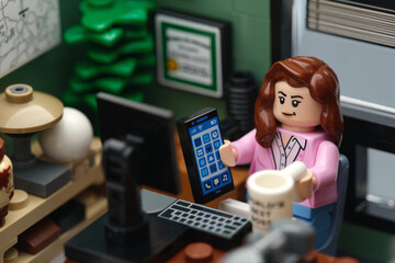 Fototapeta premium Tambov, Russian Federation - February 11, 2026 A happy Lego businesswoman minifigure sitting behind a desk in an office and holding a phone and a coffee mug. Close up.
