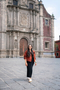 Young smiling Latina woman stands relaxed in front of a baroque church in Mexico City&rsquo;s historic center; wears black dress, rust jacket, white shoes and glasses under soft daylight.