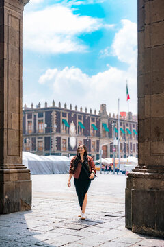 Smiling young Latina walks confidently through the historic center of Mexico City, showing joy and energy; wears black dress, rust jacket, white shoes and glasses, in bright daylight.