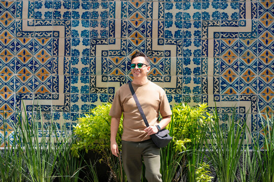 Young smiling Latino man casually in front of the vibrant tiled wall of Sanborns de los Azulejos in the historic center of Mexico City; wearing beige shirt, green pants, sunglasses and shoulder bag.