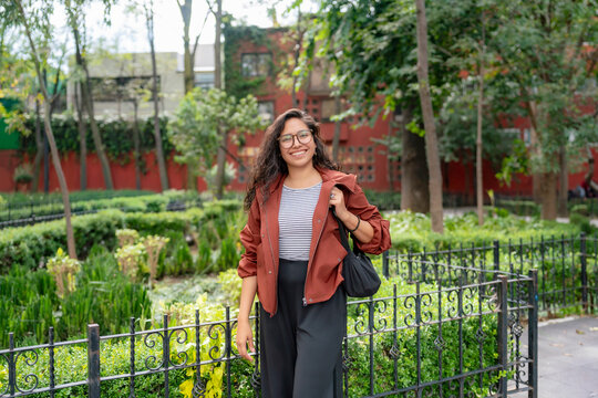Young smiling Latina woman poses with confidence and joy in a quiet garden in the center of Coyoacan, Mexico City; wearing striped shirt, rust jacket, black trousers and glasses in soft daylight