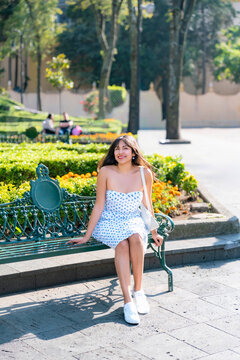 Young Latina woman smiles confidently while seated on a green bench in Centro de Coyoacan, Mexico City. Surrounded by bright flowers, wearing a white strapless dress with blue pattern and white bag.
