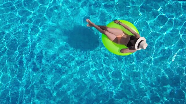 Aerial view of young woman in hat relaxing in swimming pool, beautiful girl in bikini floats on inflatable ring donut and has fun in water, tropical vacation on holiday resort, top drone view