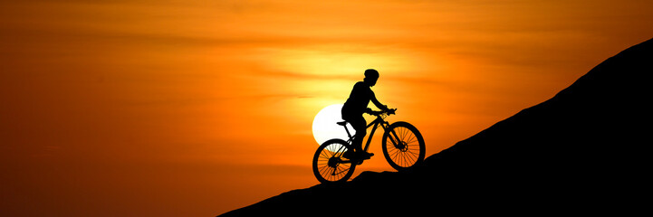 Obraz premium A cyclist silhouetted against a radiant sunset while climbing a hill. The biker is focused and determined as they navigate the incline with their bike