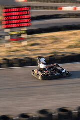 A person driving a go-kart speeds past the electronic scoreboard.