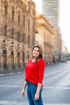 Young Latina woman smiling while crossing Eje Central Avenue in Mexico City under the morning sun, with Torre Latinoamericana behind her, wearing a red sweater and blue jeans in a calm urban scene