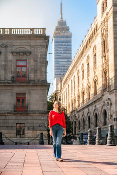 Young smiling Latina woman stands confidently in front of the MUNAL in Mexico City, with Torre Latinoamericana and Palacio Postal behind her, enjoying the sunny morning in red sweater and blue jeans