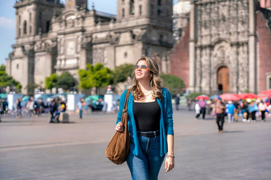 Smiling young Latina walks confidently through the Zocalo of Mexico City, in front of Cathedral Metropolitana, wearing sunglasses, blue cardigan, jeans on a sunny afternoon surrounded by urban life