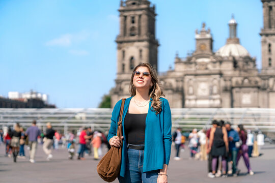 Smiling young Latina walks confidently through the Zocalo of Mexico City, in front of Cathedral Metropolitana, wearing sunglasses, blue cardigan, jeans on a sunny afternoon surrounded by urban life