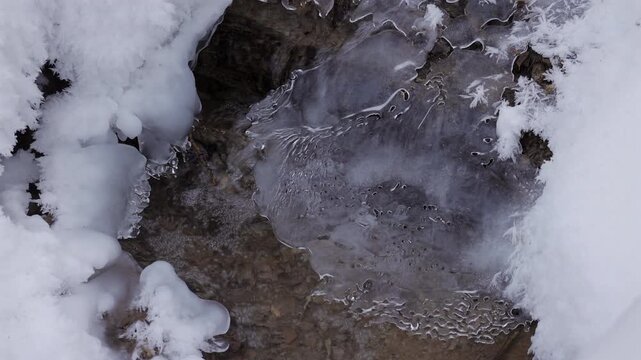 This video captures slow motion of a beautiful icy landscape where a stream flows beneath snow and ice, showcasing the intricate shapes and textures of winter in nature.