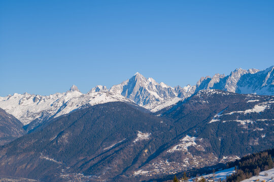 The dramatic summit of Aiguille Verte rises sharply above a winter landscape of forested slopes and scattered snowfields under a clear blue sky in the French Alps.