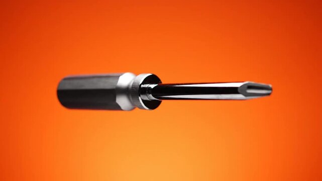 Close-up of a screwdriver rotating against a vibrant orange background, showcasing its metallic handle and tip.