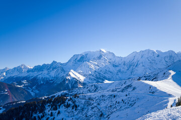 Fototapeta premium Vast panoramic view of the Mont Blanc massif with rugged, snow-covered peaks and rolling alpine slopes beneath a vibrant blue sky. Crisp winter light enhances the dramatic mountain landscape and