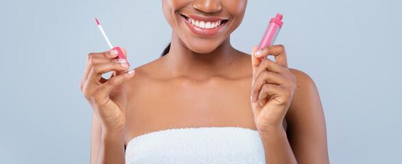 A woman stands smiling while holding a lip gloss in one hand and a lip applicator in the other hand. She appears happy and is in a bright setting, suitable for a beauty promotion. © Prostock-studio