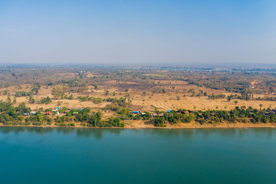 Aerial view of cultivated fields and scattered rural houses bordering the Mekong River near Stoeng Treng, Cambodia. The golden landscape stretches beneath a clear blue sky, with tranquil water in the