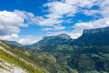 Fototapeta premium Expansive view of dramatic limestone cliffs and forested slopes in the Chartreuse Massif, with scattered clouds and vibrant green valleys below. The scene highlights the rugged terrain and natural