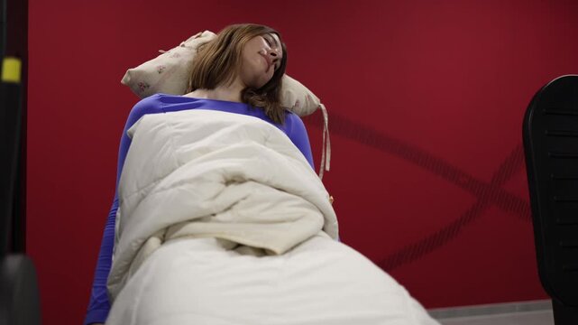 Funny woman appears asleep in gym chair wrapped in blanket while fitness machine keeps moving around her playful training paradox lazy workout humor red wall background