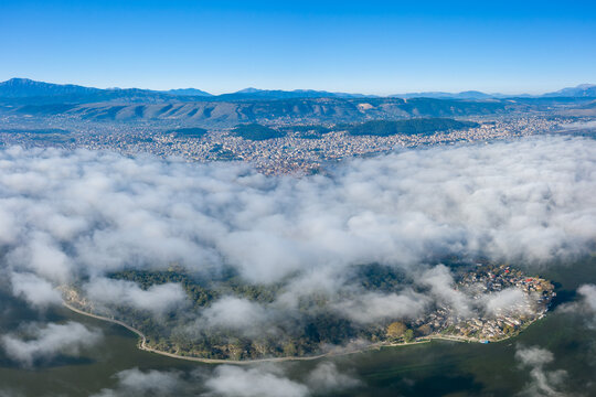 Aerial panorama shows low clouds drifting above the island village in Lake Ioannina, with the city of Ioannina and the Pindus mountains visible in the distance. Bright daylight and crisp blue sky