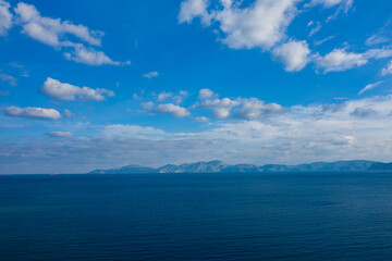 Fototapeta premium Vast blue ocean stretches toward a hazy mountain range on the horizon under a sky filled with scattered clouds. The tranquil scene captures natural gradients, open space, and a sense of calm.