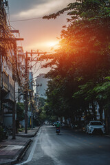 Typical Bangkok street at sunset with tropical trees and power lines, Cozy urban road in Thai city at dusk with greenery, motorcycles and cables overhead, Thailand