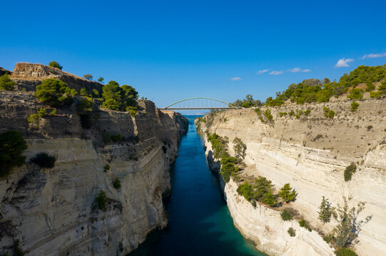 Clear blue water flows through the narrow Corinth Canal, bordered by steep, sunlit limestone walls topped with green pines. An arched bridge spans the dramatic gorge beneath a vibrant sky, creating a