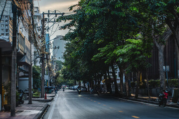 Typical Bangkok street at sunset with tropical trees and power lines, Cozy urban road in Thai city at dusk with greenery, motorcycles and cables overhead, Thailand