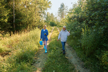 Fototapeta premium Two people walk on a dirt path through green trees while collecting plants in the afternoon sun