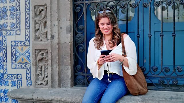 Young Latina woman sits smiling sending messages at iconic tiled wall of Casa de los Azulejos, Sanborns in the historic center of Mexico City, with natural daylight with vibrant ceramic patterns