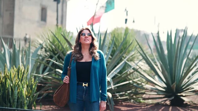 Smiling young Latina woman walks confidently beside agave plants near the Catedral Metropolitana in the Zocalo of Mexico City, wearing jeans, sunglasses, a brown bag and cardigan under bright daylight