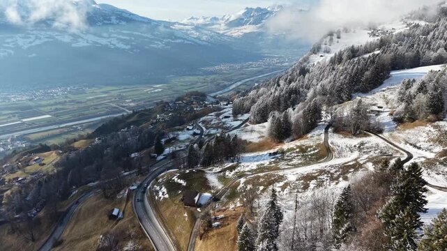 Drone aerial view along a mountain road with moving cars, showing snowy patches and clear asphalt in a scenic alpine landscape.
