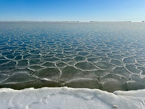 Polygonal ice formations on frozen Baltic Sea surface near snowy shoreline, natural winter patterns created by freezing water under clear sky.