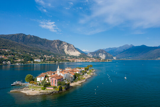 Expansive aerial perspective of Isola dei Pescatori, with its colorful buildings, church tower, and lush greenery set against the deep blue waters of Lake Maggiore. Alpine mountains and lakeside